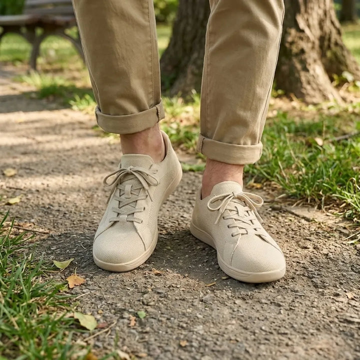 Beige knit sneakers worn with rolled khaki pants on a park path beside grass and fallen leaves.