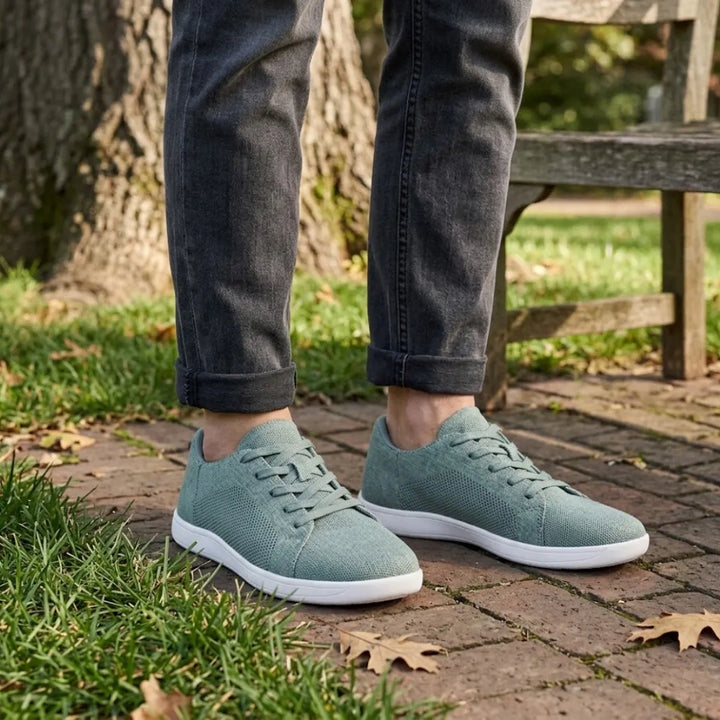 Person wearing sage green knit sneakers with white soles and rolled gray jeans, standing on a brick path near a park bench. 29