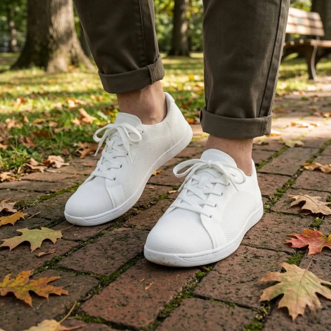 Person wearing all-white knit sneakers with olive pants rolled at the ankle, walking on a brick path covered in autumn leaves. 38