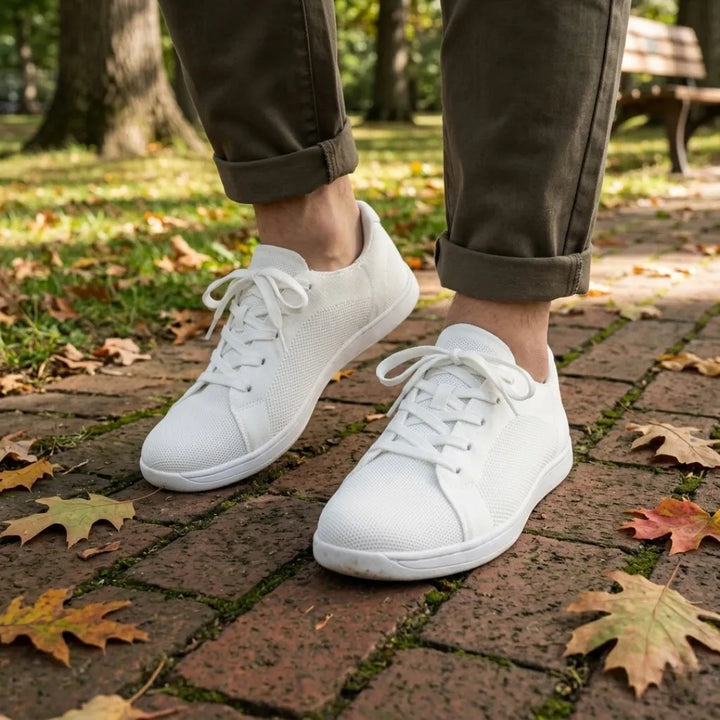 Person wearing all-white knit sneakers with olive pants rolled at the ankle, walking on a brick path covered in autumn leaves. 38