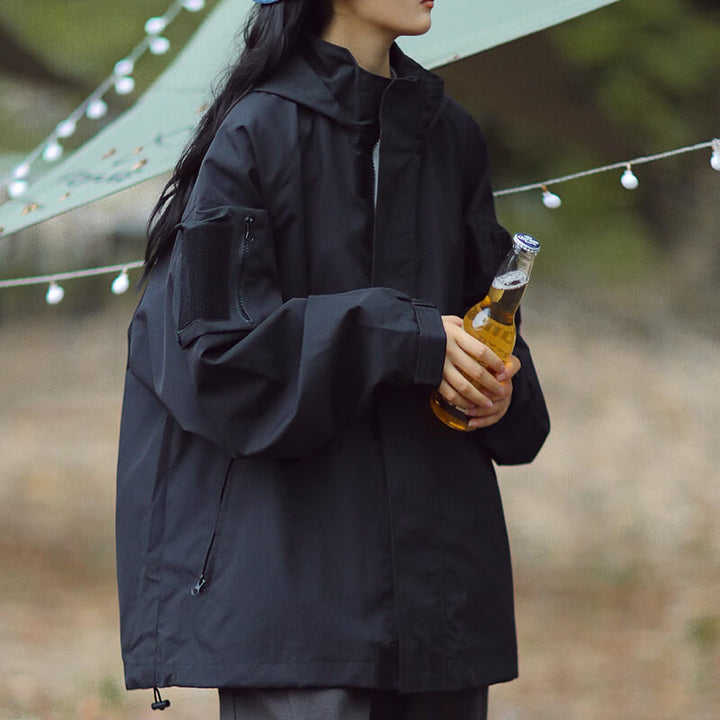 Black hooded jacket with a zippered sleeve pocket, shown on a model outdoors.