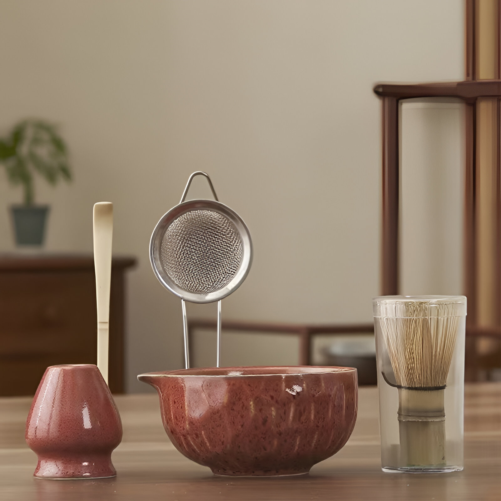 Another view of a glossy reddish-brown matcha set with pouring bowl, whisk holder, mesh sifter, bamboo scoop and whisk in a clear tube.