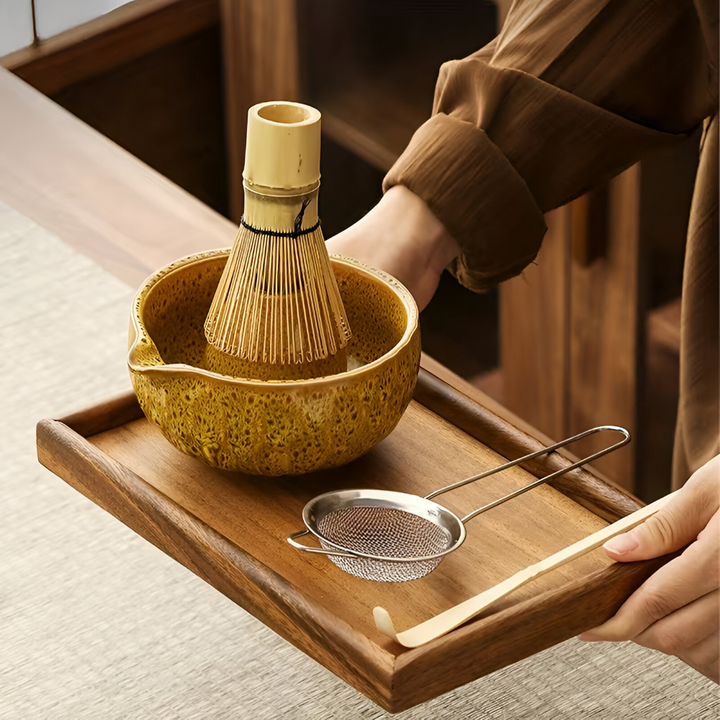 Hands carry a wooden tray holding a mustard-glazed matcha bowl with a bamboo whisk, a mesh sifter and a bamboo scoop.