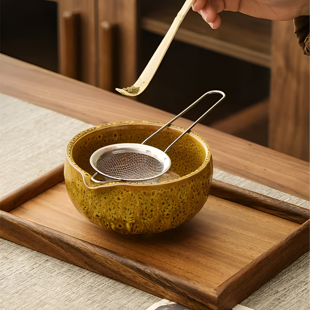 Detail of a bamboo scoop holding matcha powder above a mustard-glazed bowl with a mesh sifter on a wooden tray.