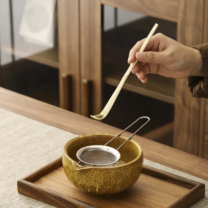 Detail of a hand holding a bamboo scoop with matcha powder over a glazed bowl fitted with a steel mesh sifter.