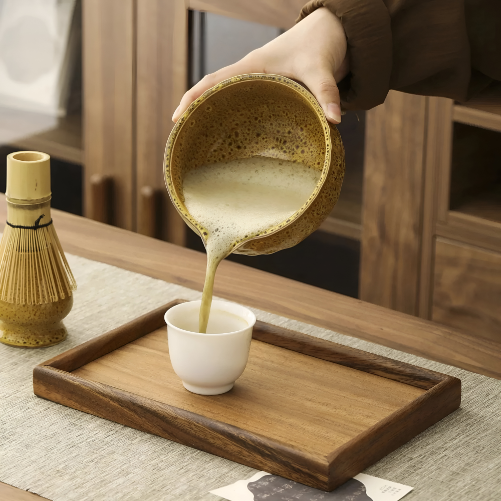Frothy matcha is poured from the spouted ceramic bowl into a small white cup on a wooden tray, with a whisk on its holder nearby.