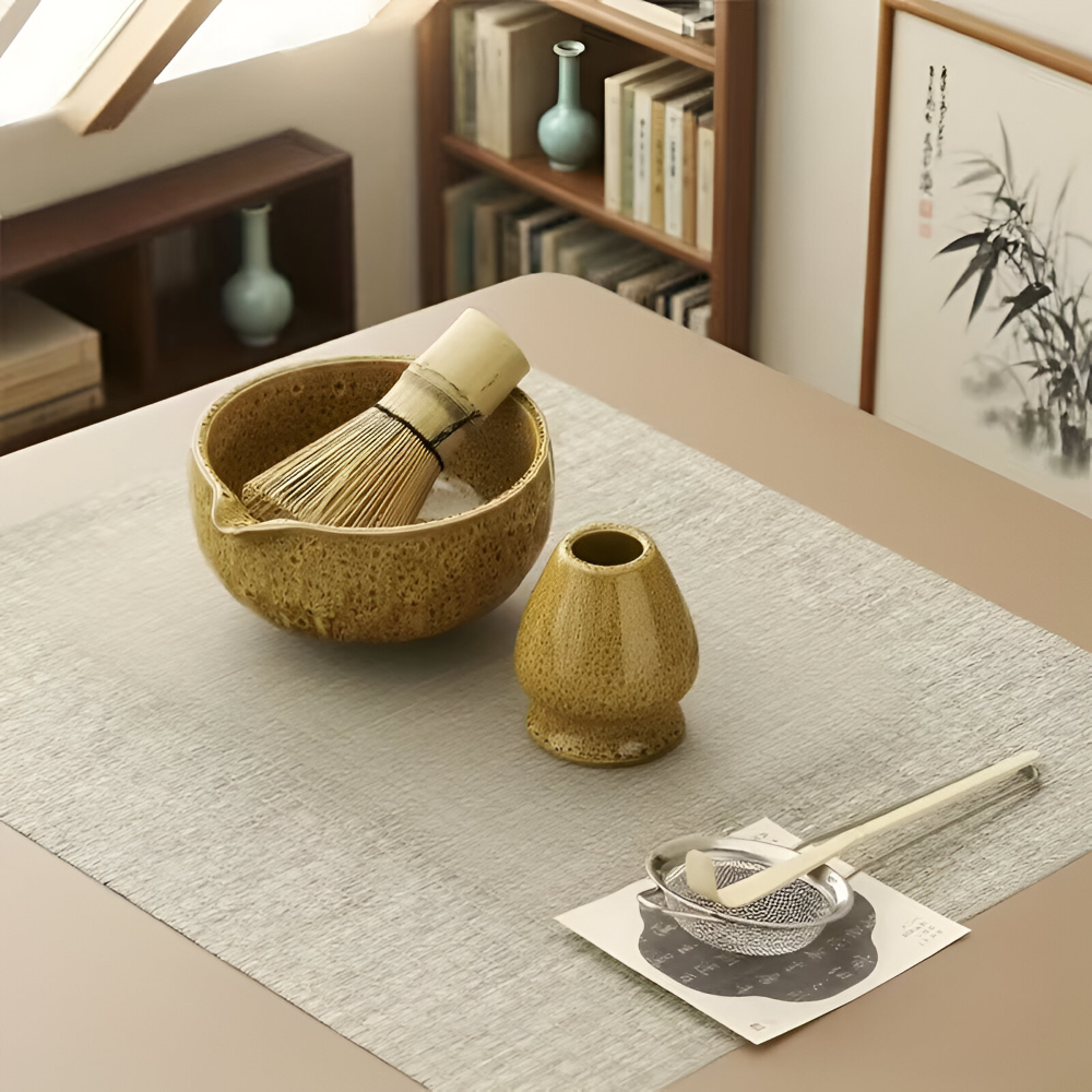 Overhead view of the matcha set on a linen runner with bowl, bamboo whisk, ceramic holder, sifter and scoop on a paper mat.