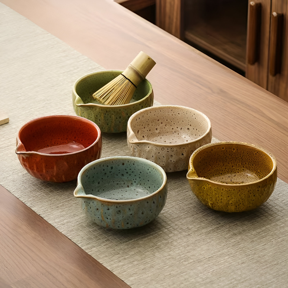 Five spouted ceramic matcha bowls in red, blue-green, cream, olive green and mustard glazes, one holding a bamboo whisk.