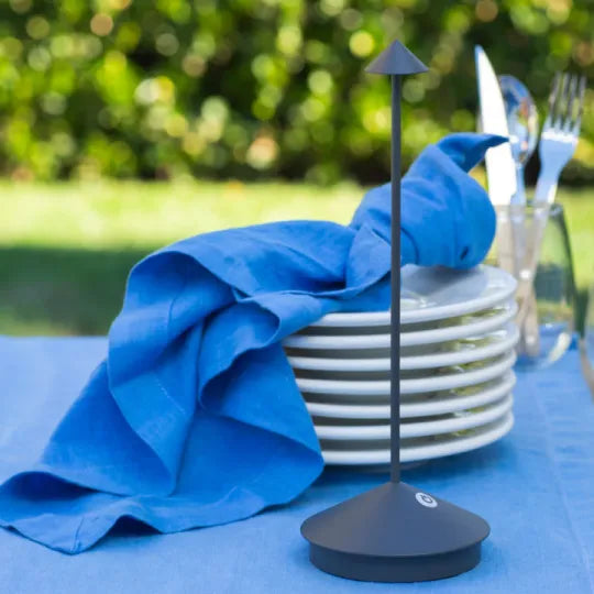 Dark gray lamp on a blue tablecloth outdoors, next to a stack of white plates and a blue napkin.