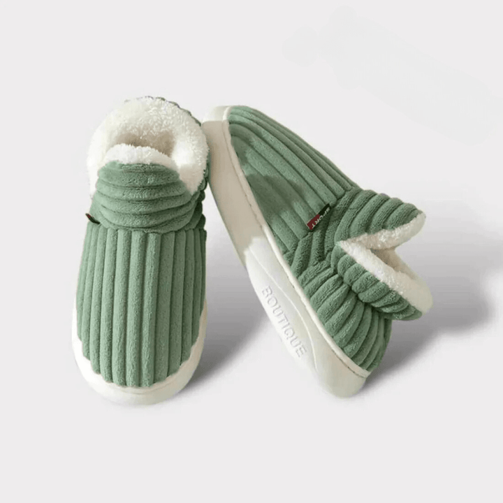 Pair of green ribbed slippers on a plain light background, one standing upright to show the thick white sole.