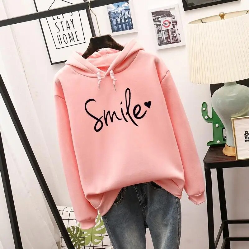 Light pink version of the pullover hoodie with black script graphic and heart, hung on a hanger over jeans.