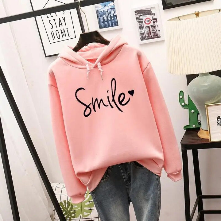 Light pink version of the pullover hoodie with black script graphic and heart, hung on a hanger over jeans.