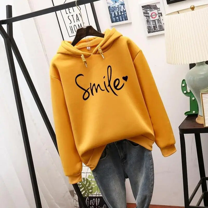 Mustard yellow hoodie with drawstring hood and black script graphic with heart, hanging above blue jeans.