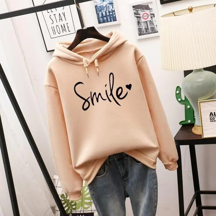 Beige drawstring hoodie with a black handwritten graphic and small heart, hanging on a hanger above blue jeans.
