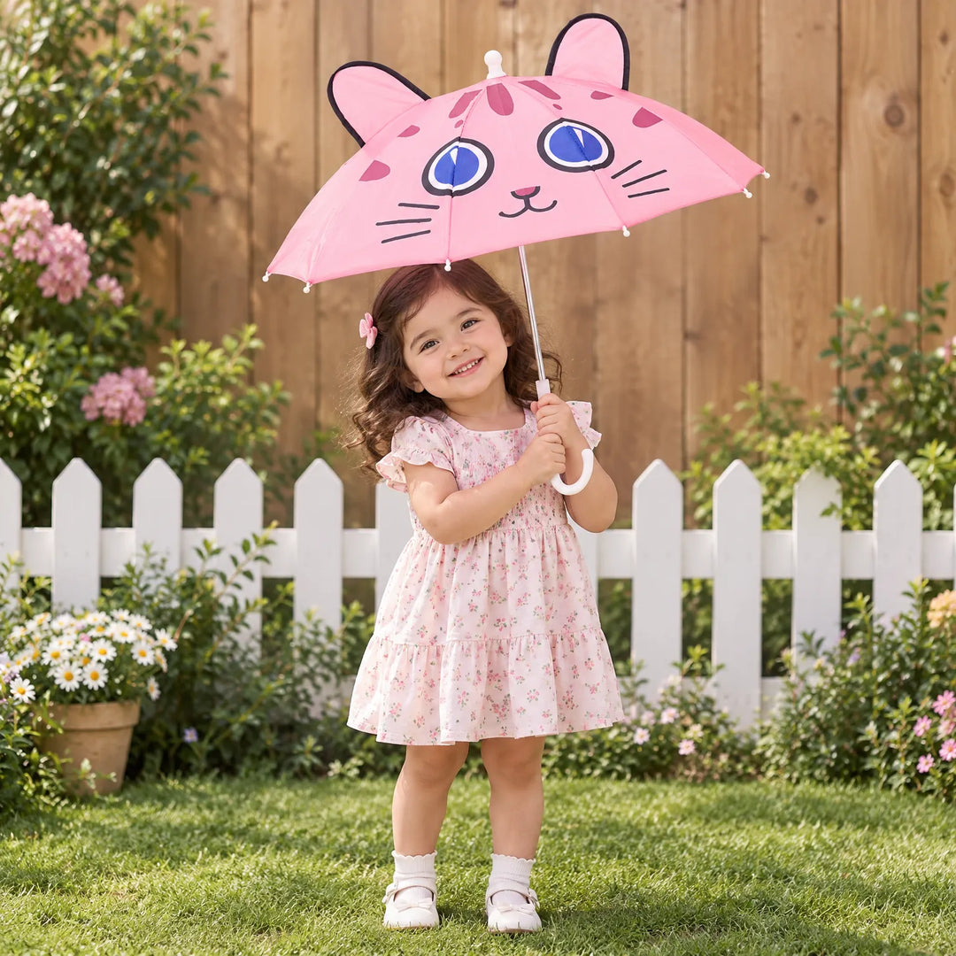 Smiling toddler in a floral dress holds the pink cat-face umbrella with ears over her head on a sunny garden lawn. 1