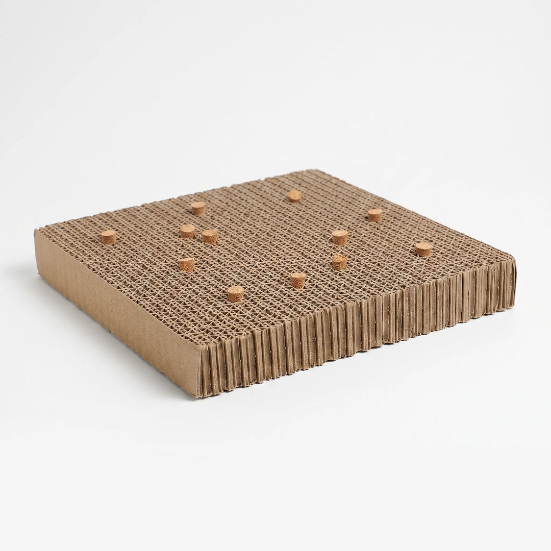Thick corrugated cardboard chew platform with small cork pieces pushed into its honeycomb top, shown at an angle on a white background. 1