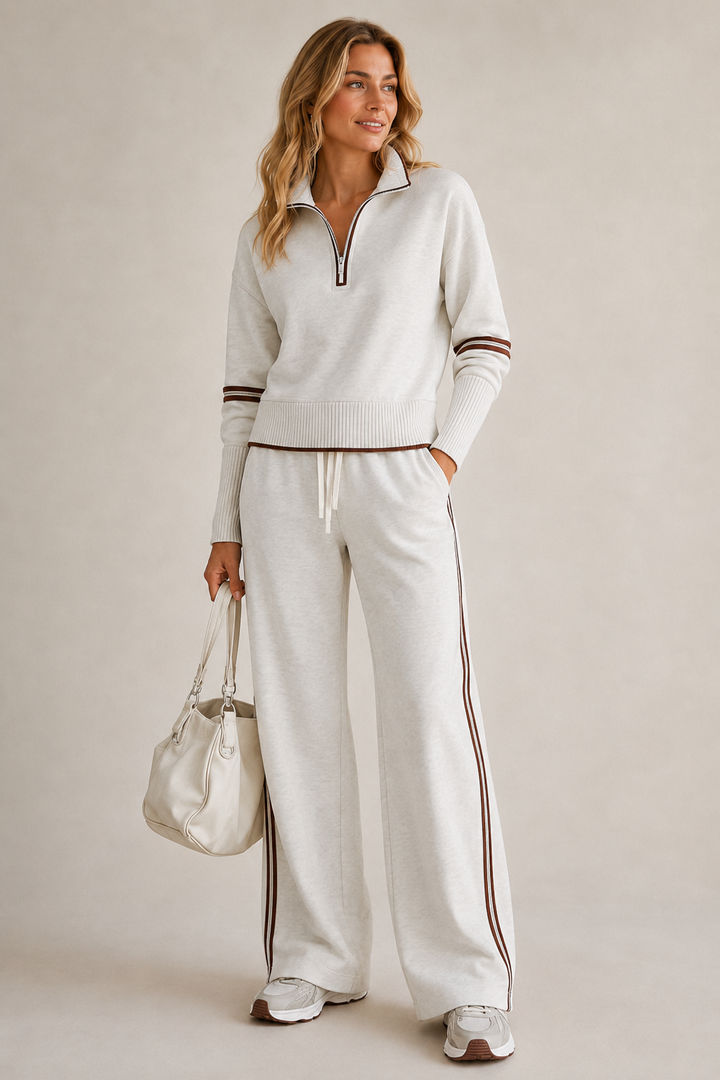 Woman in the light heather gray set with brown stripe trim, hand in pocket, holding a cream handbag, white sneakers. 1