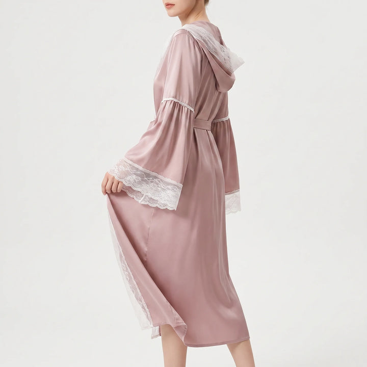 Side view of a model in the pink satin robe with its sash tied, lifting the skirt to show wide lace sleeve cuffs. 3