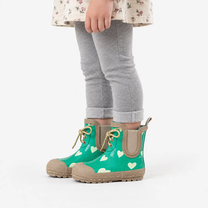 Side view of a child in gray leggings and a floral dress wearing the green heart-print boots with tan laces and toe caps. 4