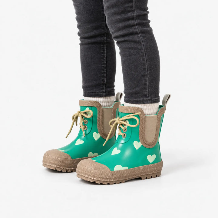 Child in dark jeans and cream socks wearing the green heart-print boots, seen from the side on a white background to show the laces. 5