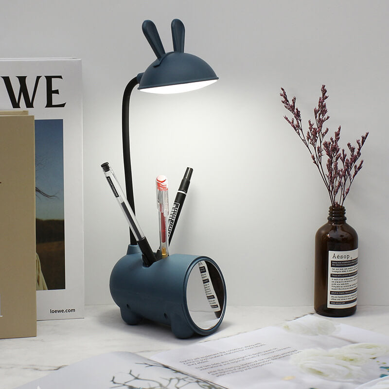 Navy bunny lamp lit on a desk, its base holding three pens, beside a dried flower vase and open book. 9