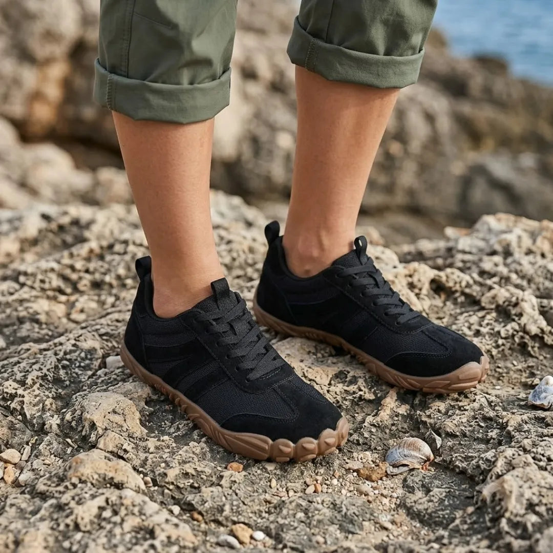 Person wearing black Lilia Women's Barefoot Shoes standing on rough coastal rocks.