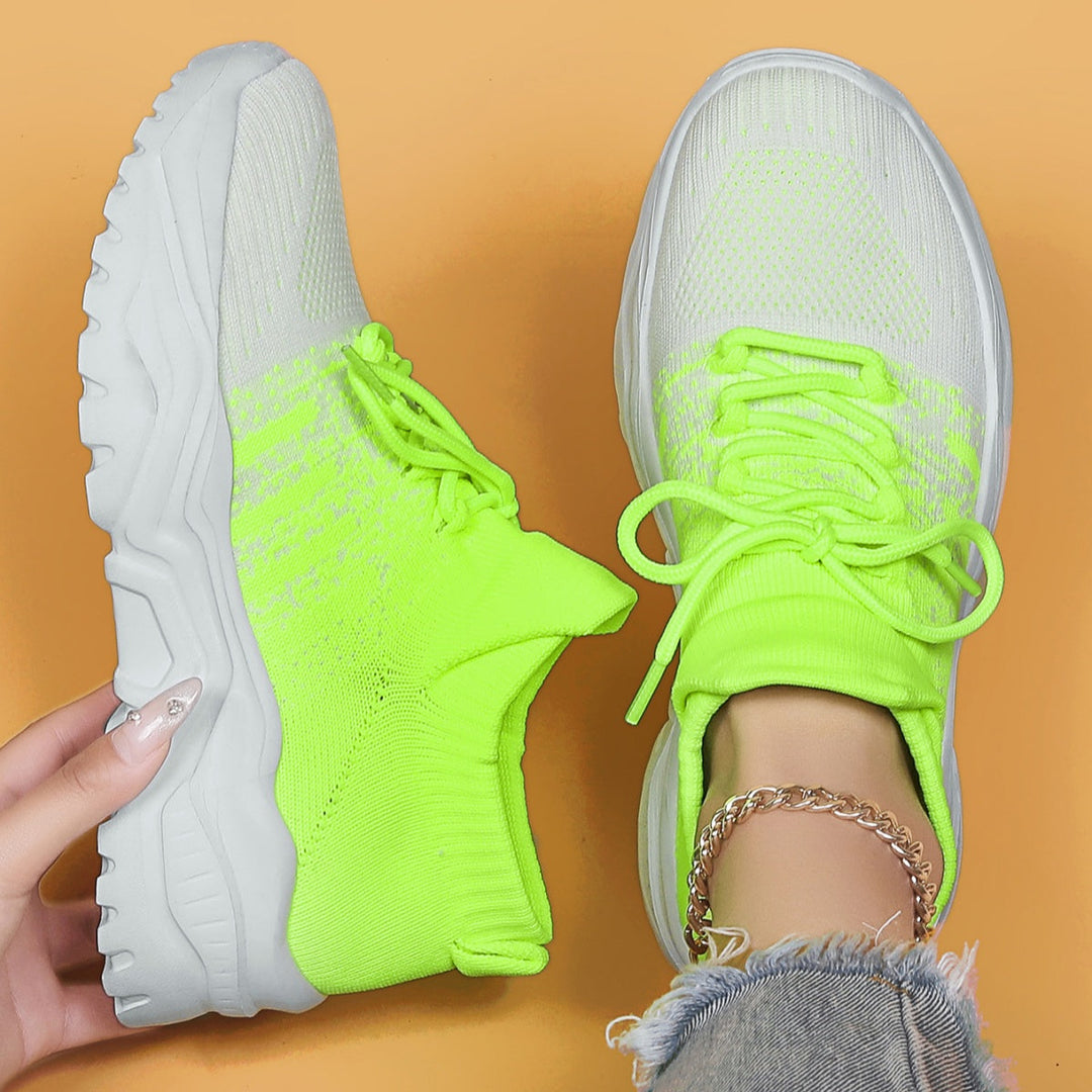 Top-down view of neon green and white knit lace-up sneakers with chunky white soles, one worn with an ankle chain, one held in hand. 21