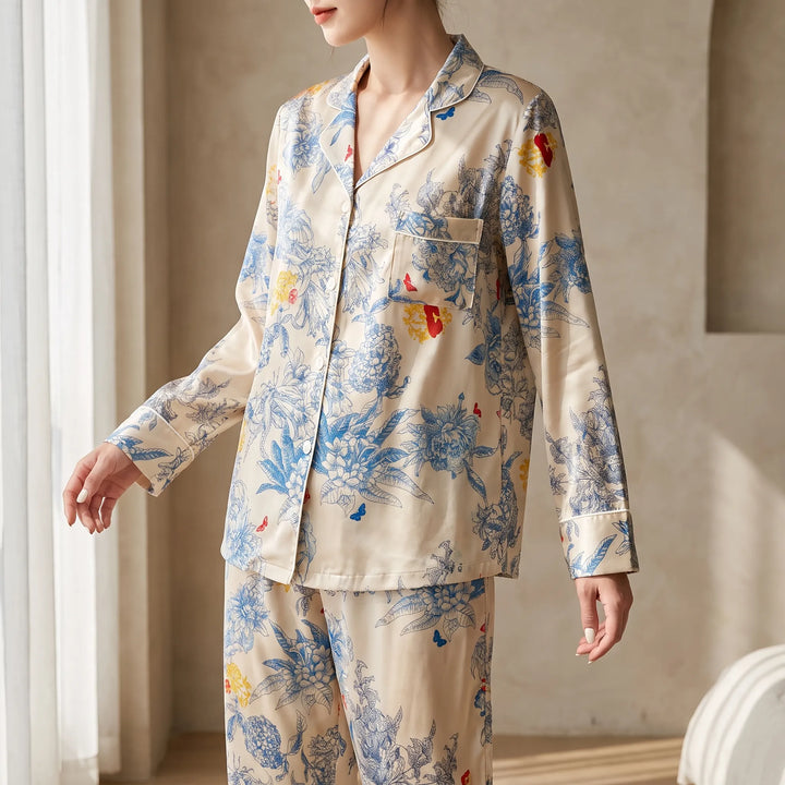 Model in a cream satin-look pajama set with blue floral print, standing beside a sunlit window, shown from the side. 1