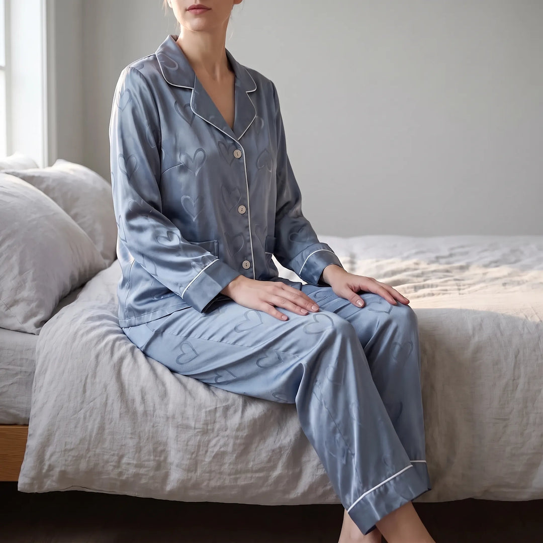 Woman sitting on the edge of a bed in blue satin pajamas with tonal heart pattern and white piping, lifestyle shot. 1