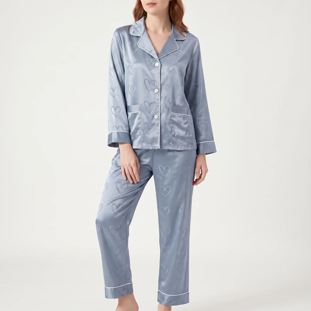 Front view of a model in the blue satin pajama set with heart jacquard, white piping and two chest pockets. 2
