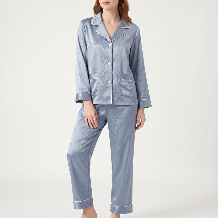 Front view of a model in the blue satin pajama set with heart jacquard, white piping and two chest pockets. 2