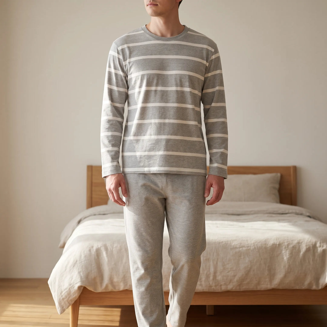 Man in gray and white striped crew-neck top and plain gray pants standing in a sunlit bedroom beside a wooden bed. 1