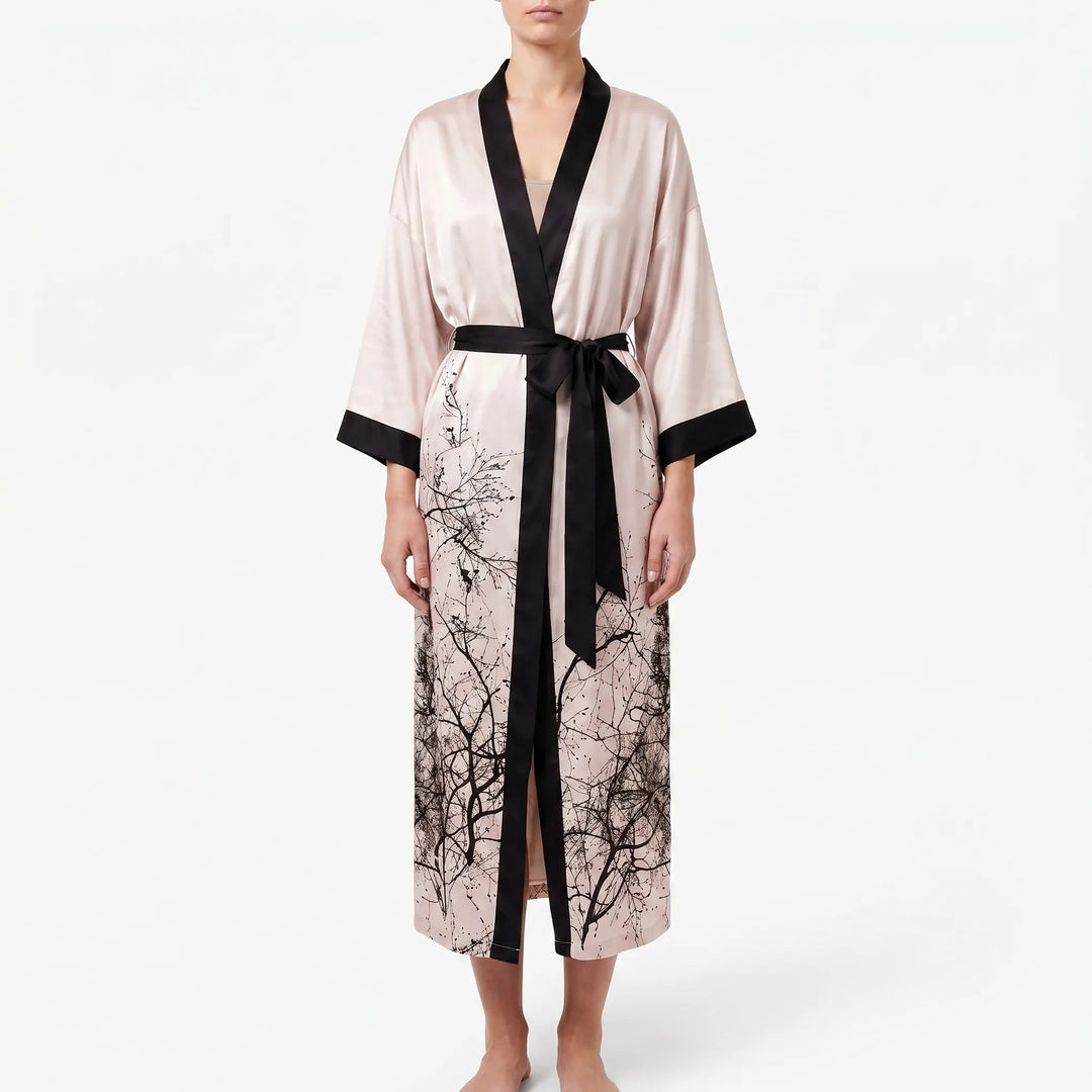 Front view of a blush satin kimono robe with black trim, belted at the waist, branch print on the hem and a front slit. 12