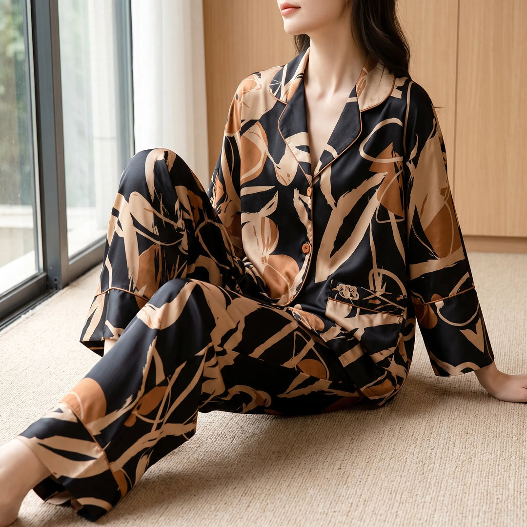 Woman sitting on a beige carpet by a window, wearing the satin pajama set in black with tan and copper abstract print. 1