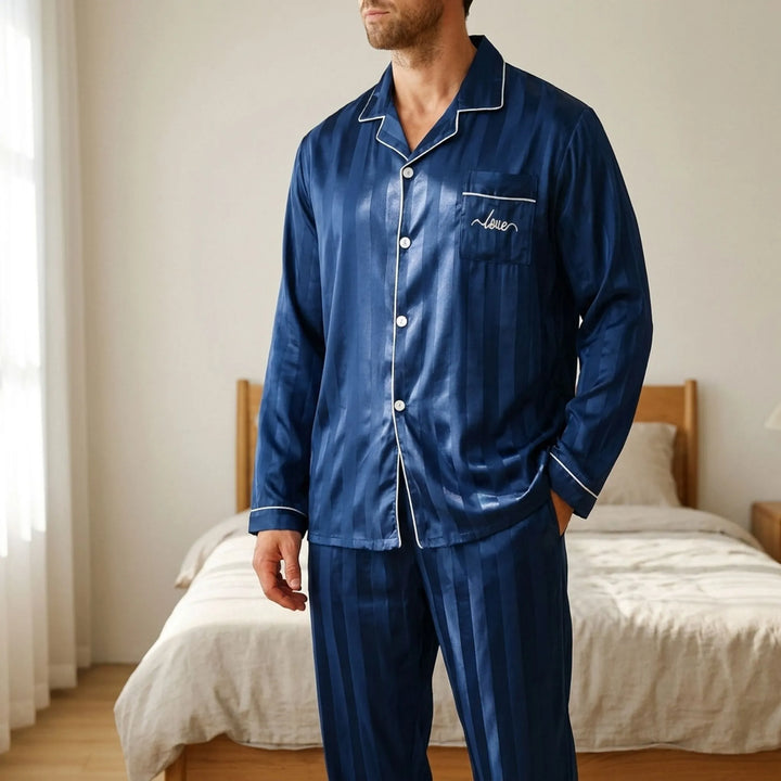 Man in navy striped satin pajama set with white piping stands in a sunlit bedroom, one hand in his pocket. 1