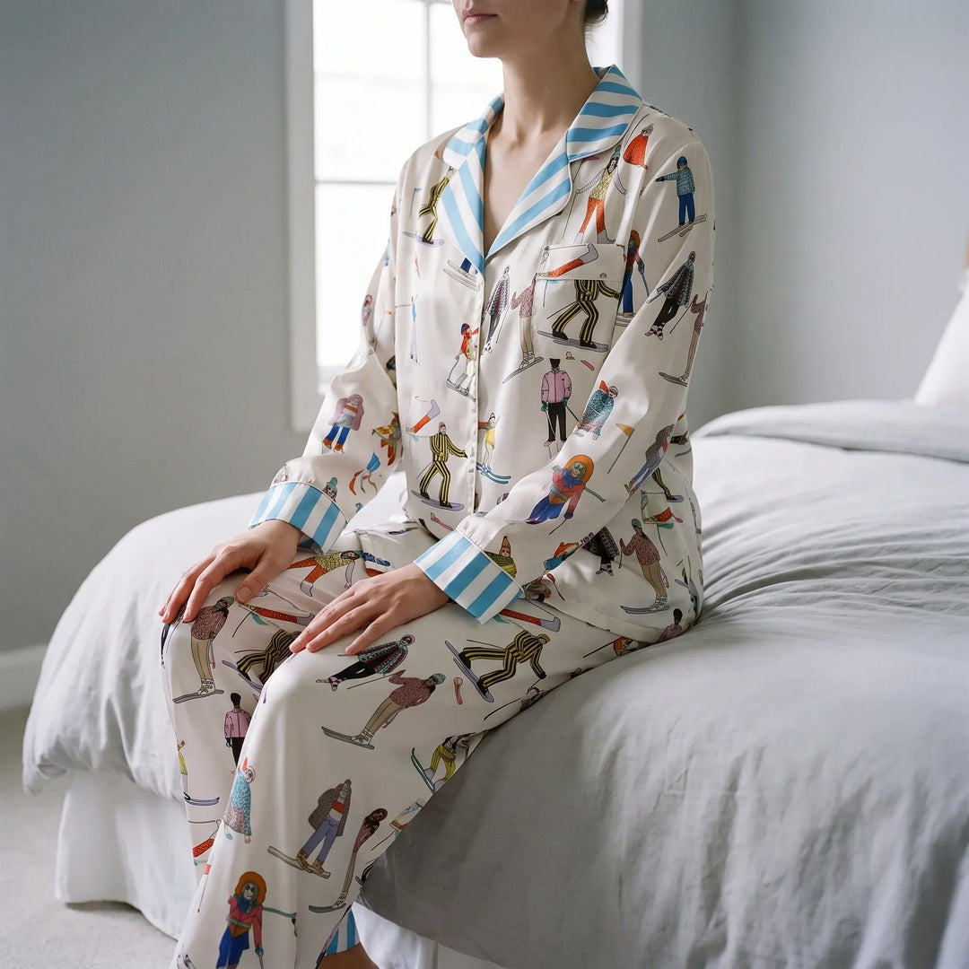 Model seated on the edge of a bed in the cream satin pajamas with skier print and blue striped collar and cuffs. 1