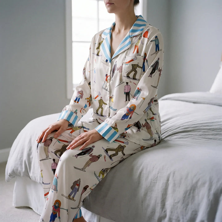 Model seated on the edge of a bed in the cream satin pajamas with skier print and blue striped collar and cuffs. 1