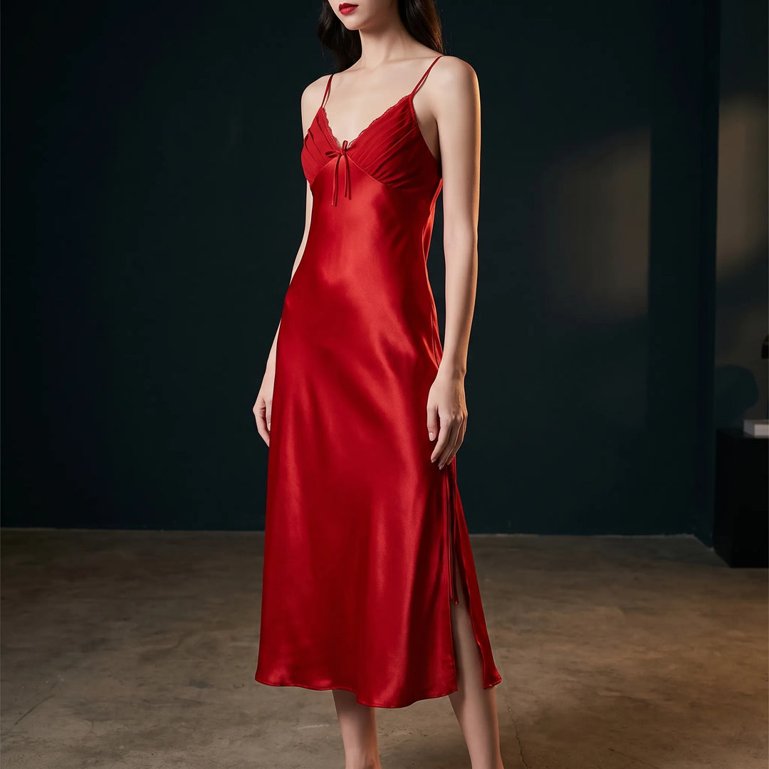 Model wears the red satin slip nightdress in a dark studio, showing the pleated lace-trimmed bodice, bow and side slit. 1