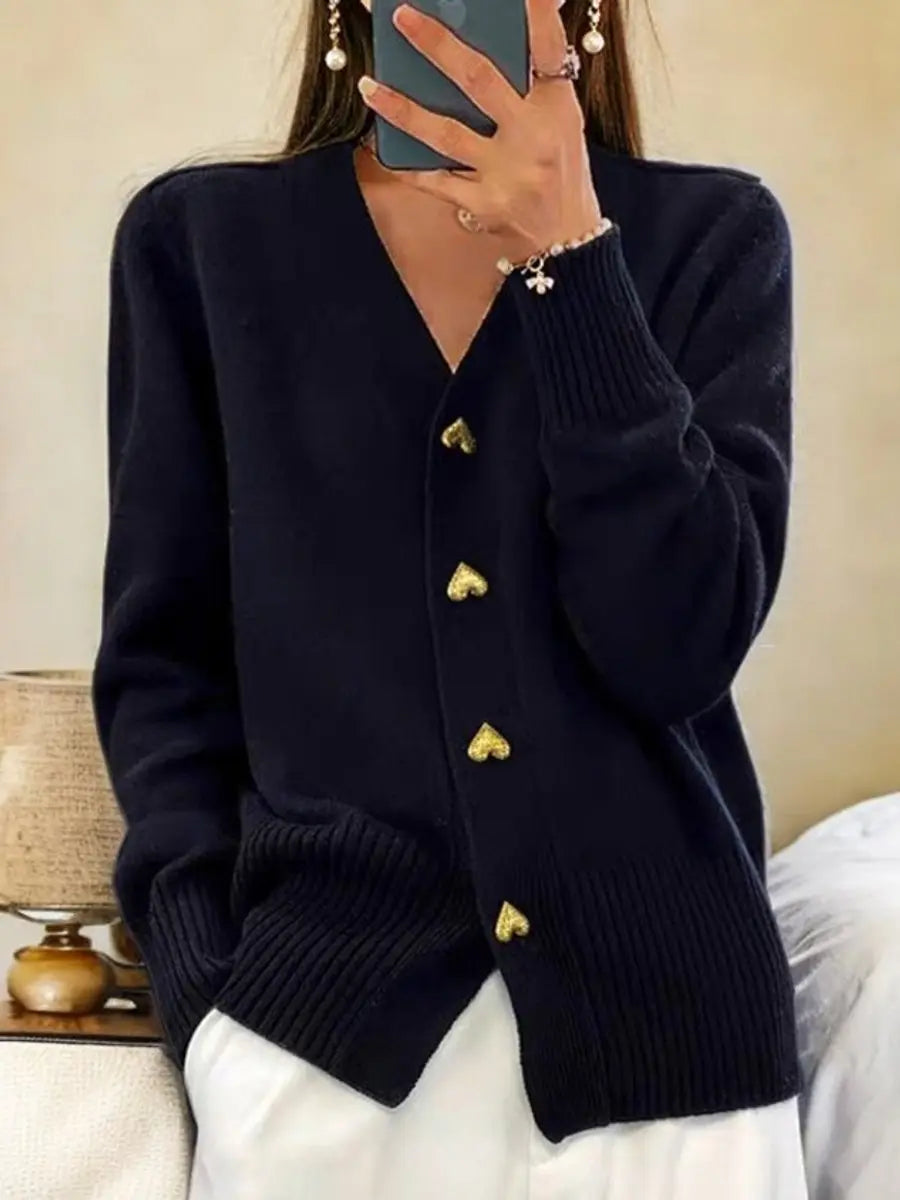 Woman taking a mirror selfie in a black wool cardigan with gold heart buttons, ribbed cuffs and hem, over white pants.