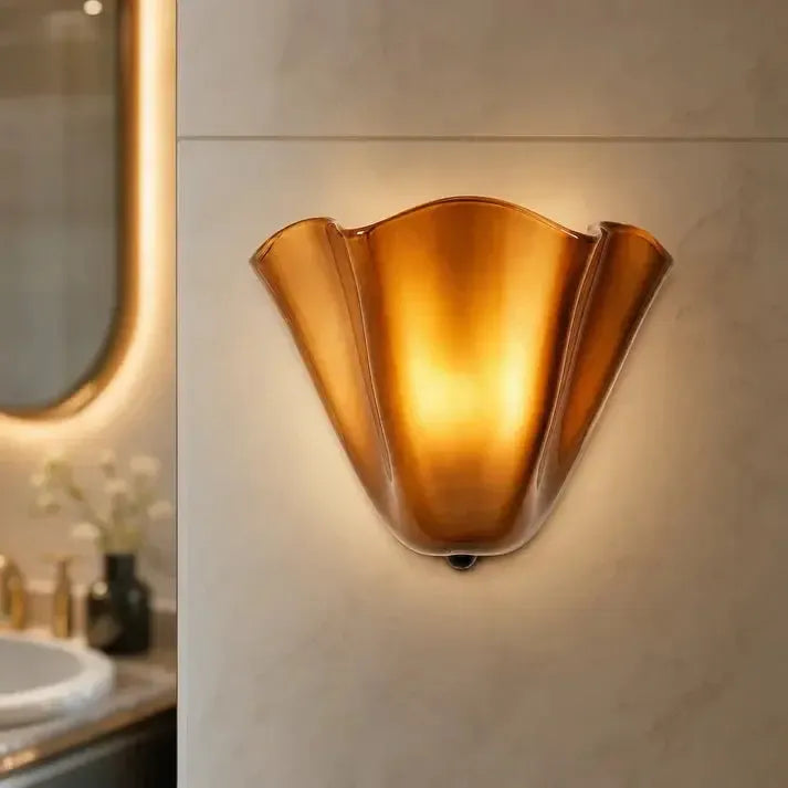 Close view of a glowing amber ruffled sconce on a marble-look bathroom wall beside a lit mirror. 10