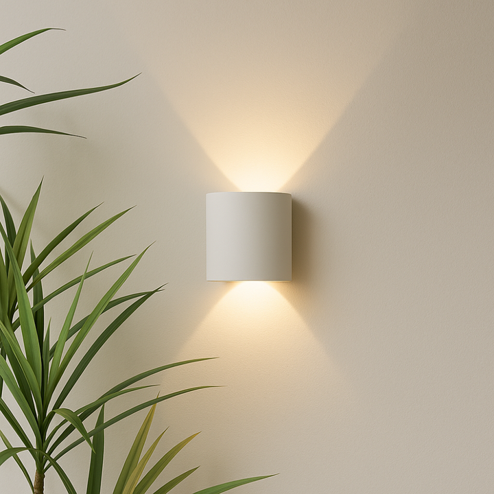 White cylindrical wall light mounted on a cream wall, glowing warm up and down, with plant leaves in the corner.