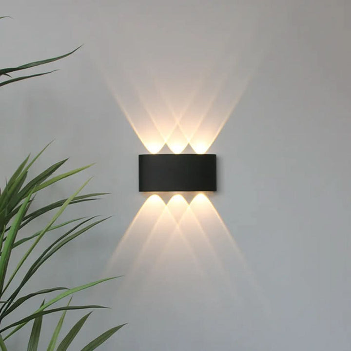 Black oval wall light detail with three beams up and three down, overlapping into diamond patterns on a gray wall.