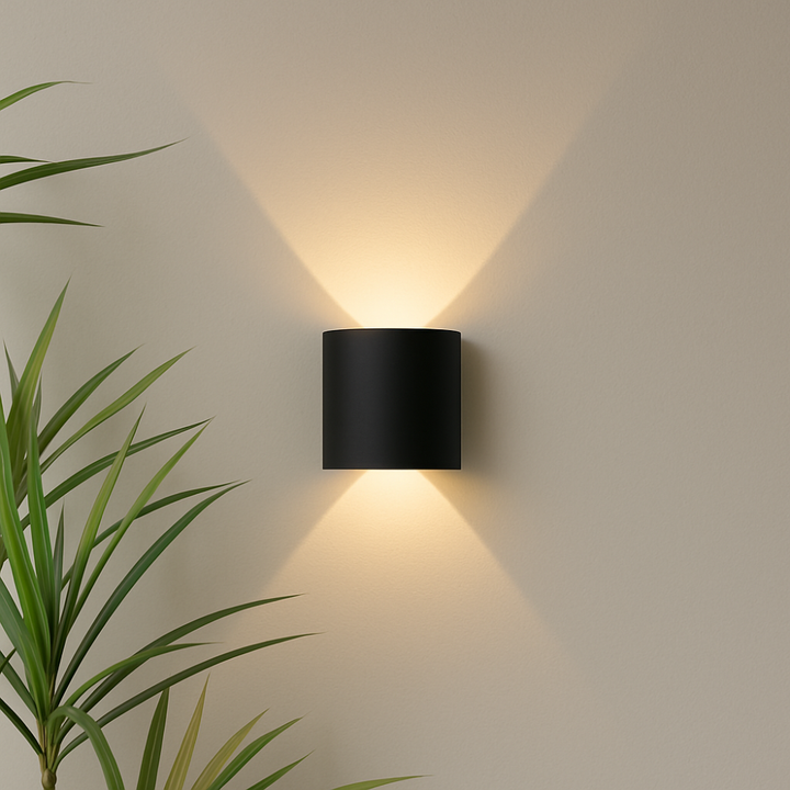 Black cylindrical wall light mounted on a beige wall, casting warm light up and down, with a leafy plant beside it.