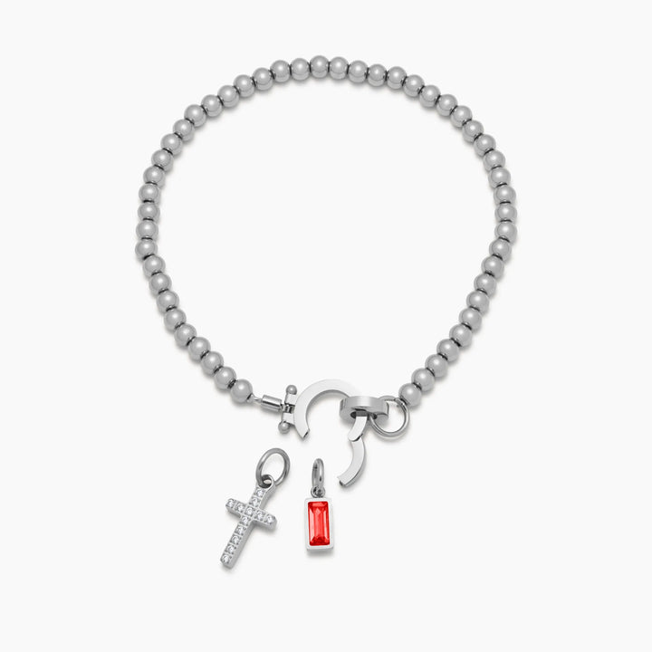 Silver-tone bead bracelet detail with the round clasp opened and the crystal cross charm and red stone charm detached below it.
