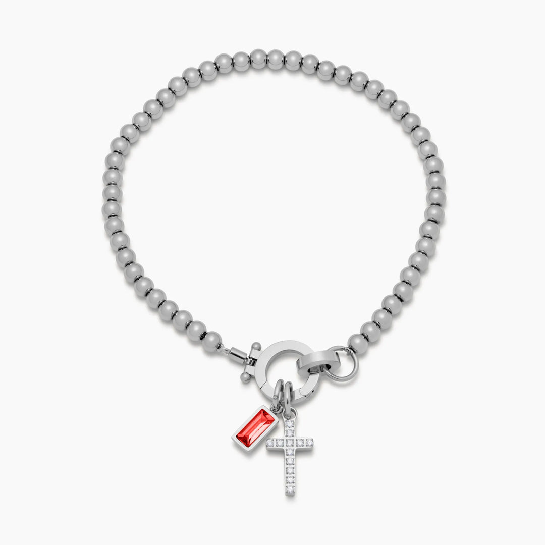 Silver-tone bead bracelet detail laid flat on white, with a red baguette stone charm and a crystal-set cross charm on the round clasp.