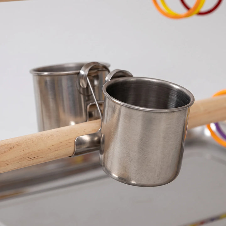 Close-up of two stainless steel feeding cups clipped onto a natural wood perch above the metal tray, with colorful rings behind. 3