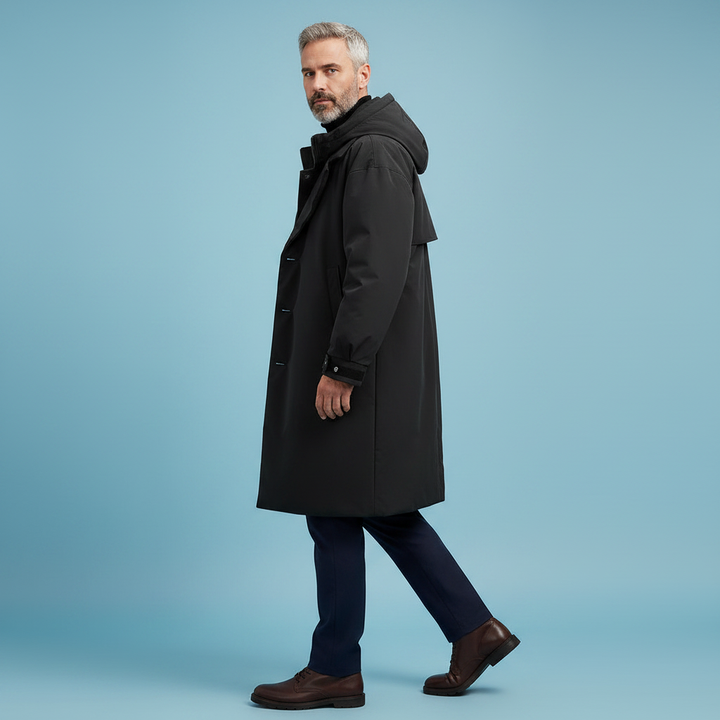 Side view of a gray-haired model walking in the long black hooded coat with navy trousers and brown leather shoes.