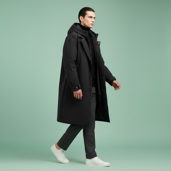 Model strides in profile wearing the long black hooded coat unzipped over a dark turtleneck, gray trousers and white sneakers.
