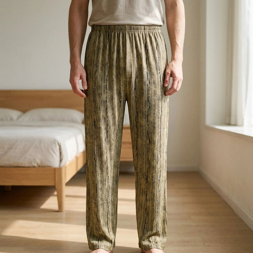 Man stands barefoot in a sunlit bedroom wearing loose olive and black textured striped pajama bottoms with an elastic waistband. 1