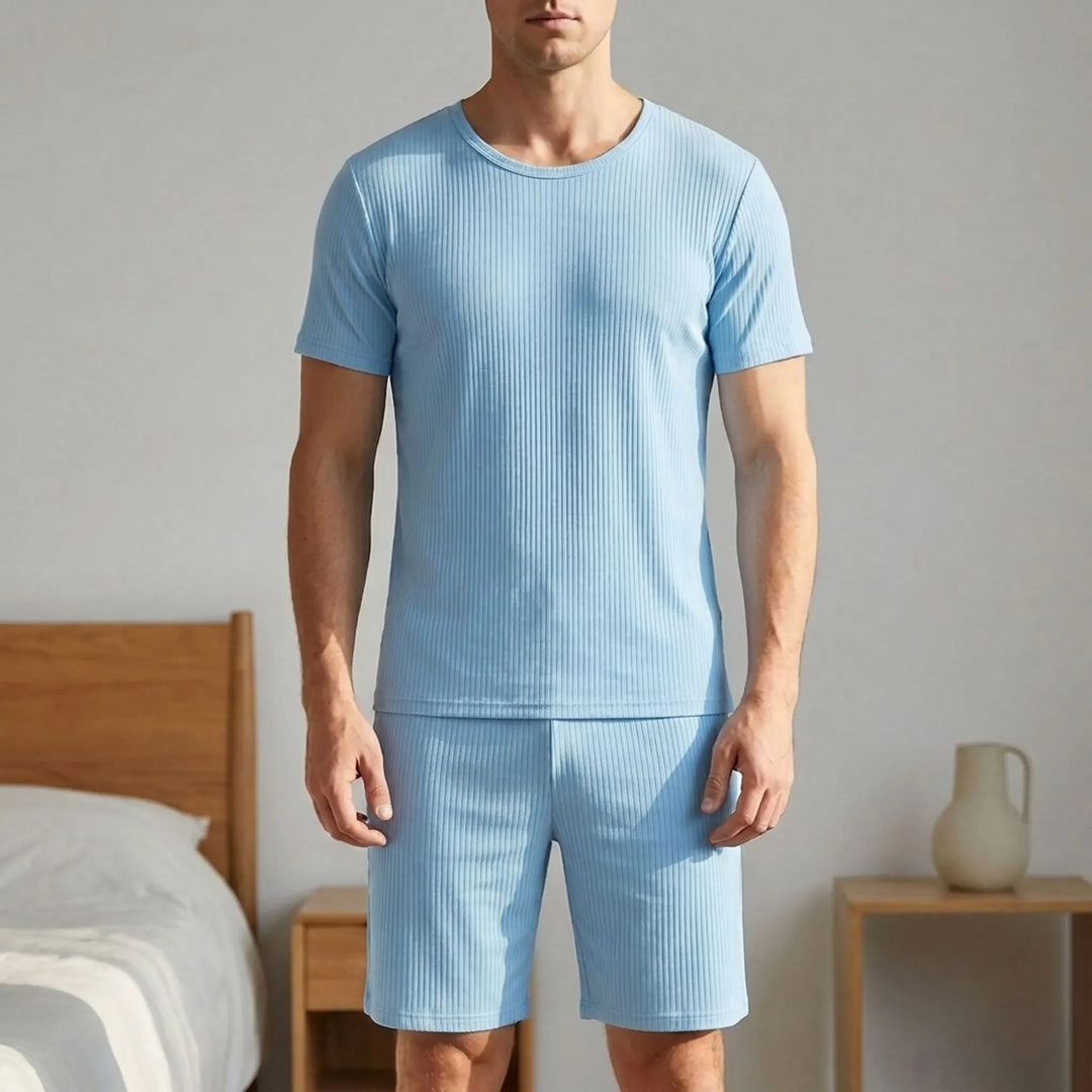 Man in a light blue ribbed short-sleeve tee and shorts standing in a bedroom beside a wooden bed and nightstand. 1